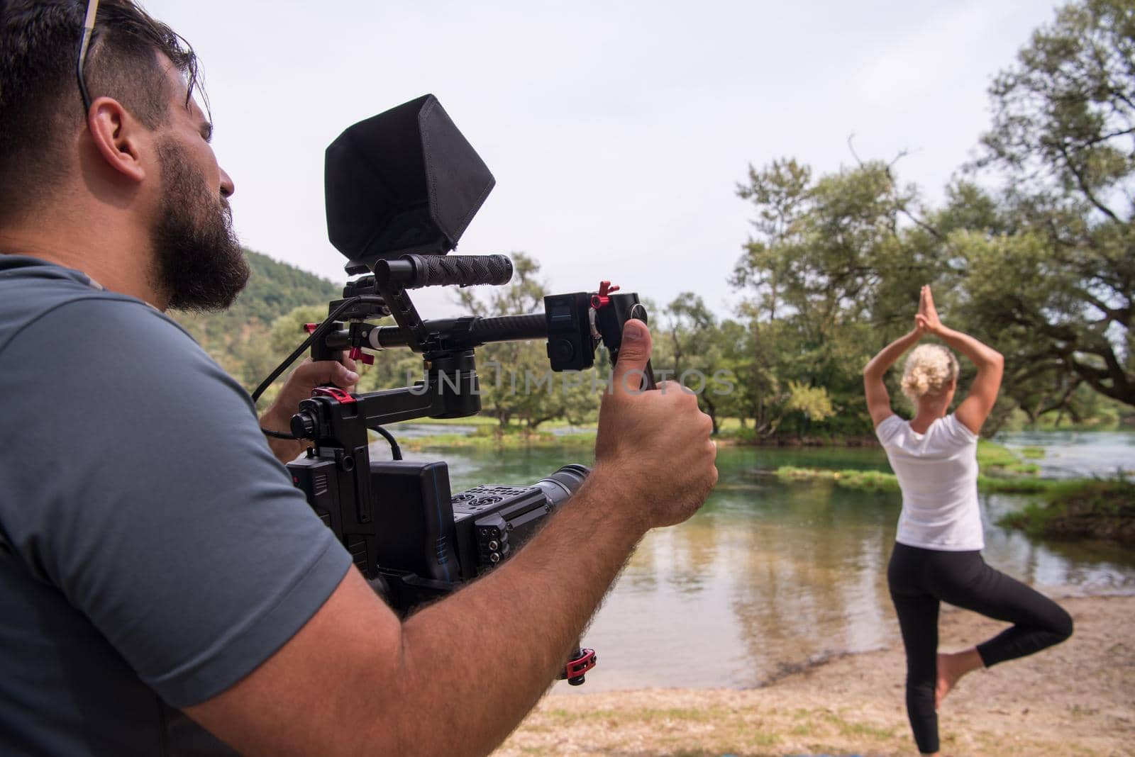 young videographer with gimball video slr rocording while healthy woman doing yoga exercise in the beautiful nature on the bank of the river