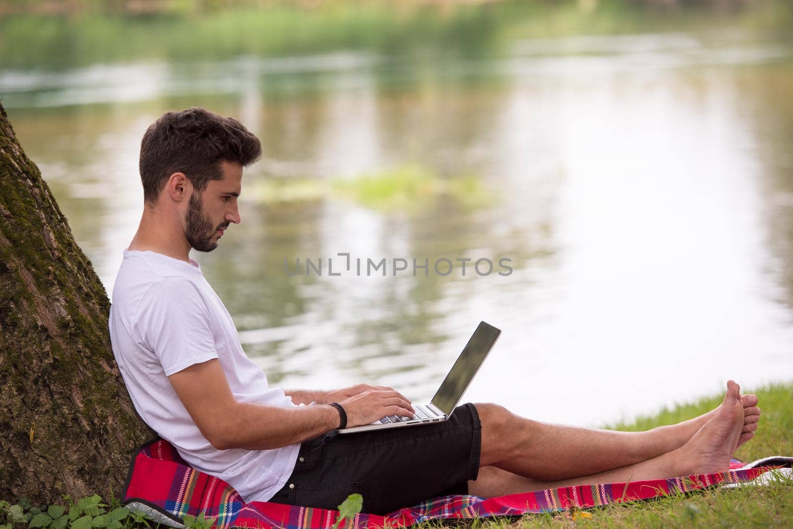 A young freelancer using a laptop computer while working in beautiful nature under the tree on the bank of the river