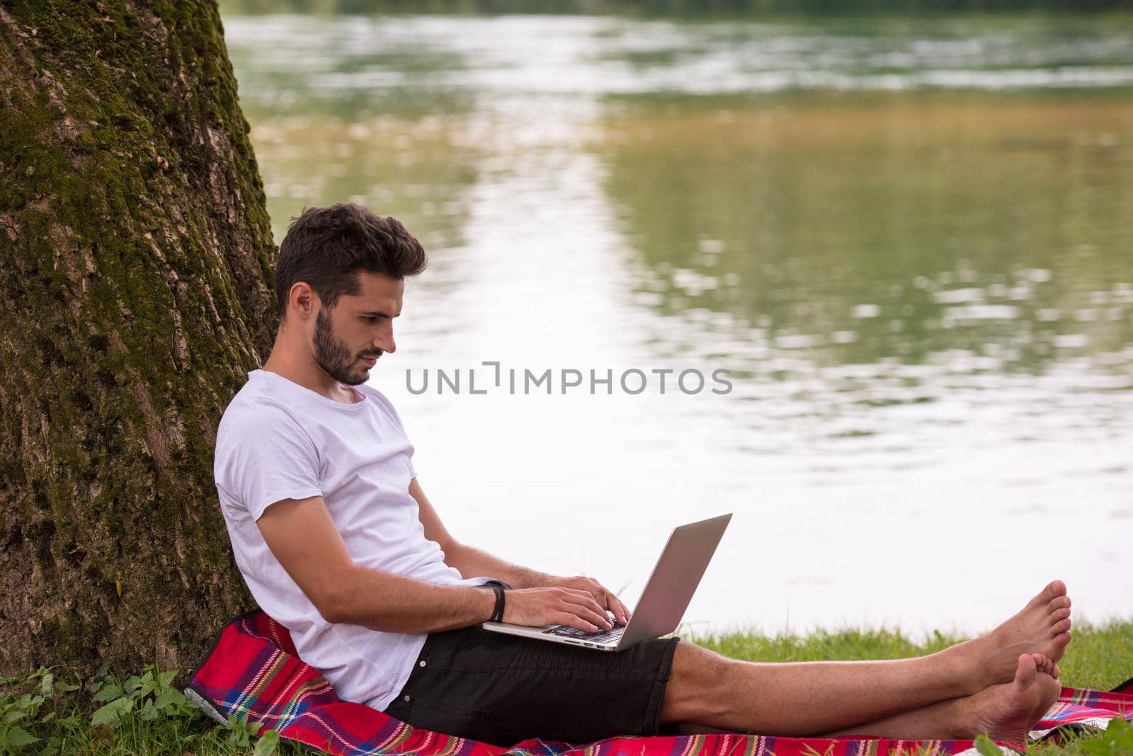 A young freelancer using a laptop computer while working in beautiful nature under the tree on the bank of the river