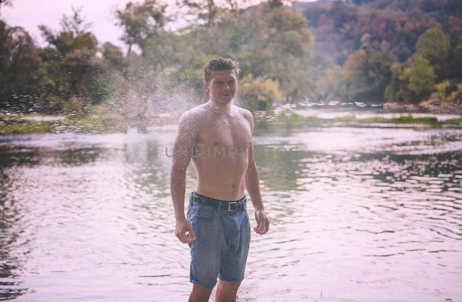 a young strong man with a bare torso having fun splashing water at sunset on the river