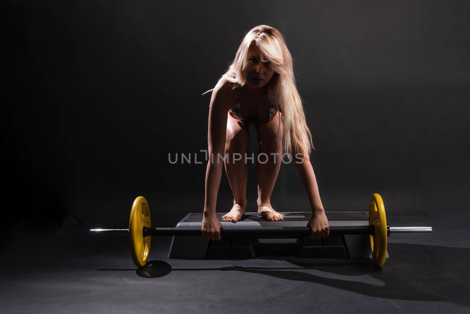 Fitness with a barbell girl blonde athlete on a black background smiles young body, barbell woman strength weight, bodybuilding dark. practice squat sportswear