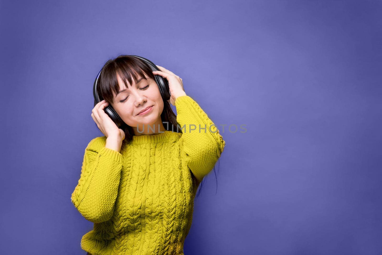 A brunette woman in a yellow sweater is listening to music with headphones. Demonstrating the colors of 2022 - Very Peri. Changing colors of the year concept. Illuminating and very pari.