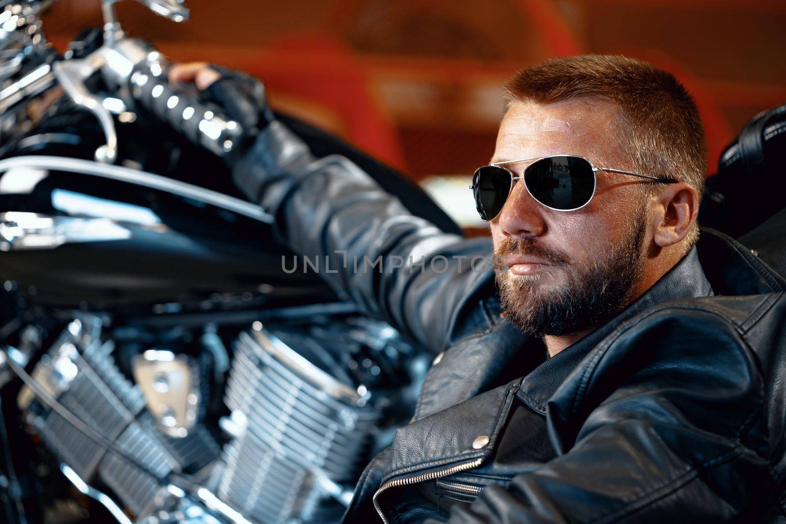 Cool man biker in sunglasses sitting near his motocycle in dark building