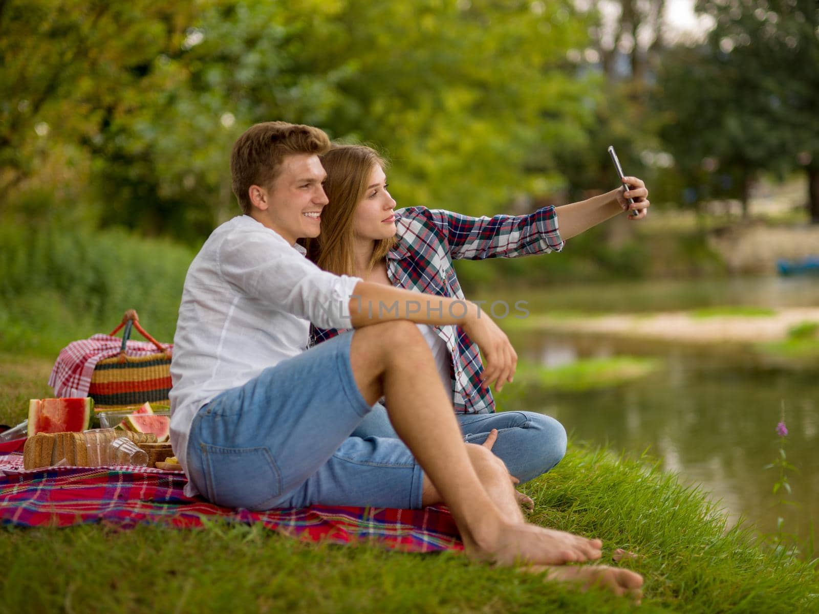 Couple in love taking a selfie by mobile phone while enjoying picnic time drink and food in beautiful nature on the river bank