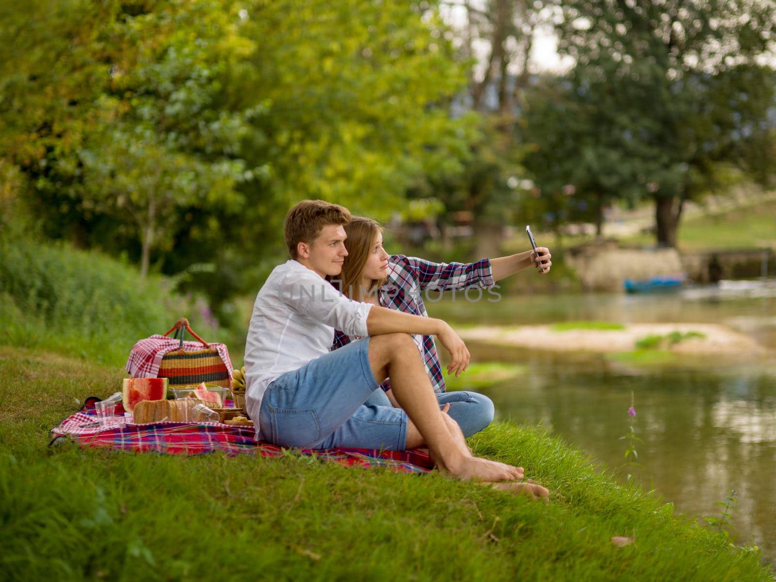 Couple in love taking a selfie by mobile phone while enjoying picnic time drink and food in beautiful nature on the river bank