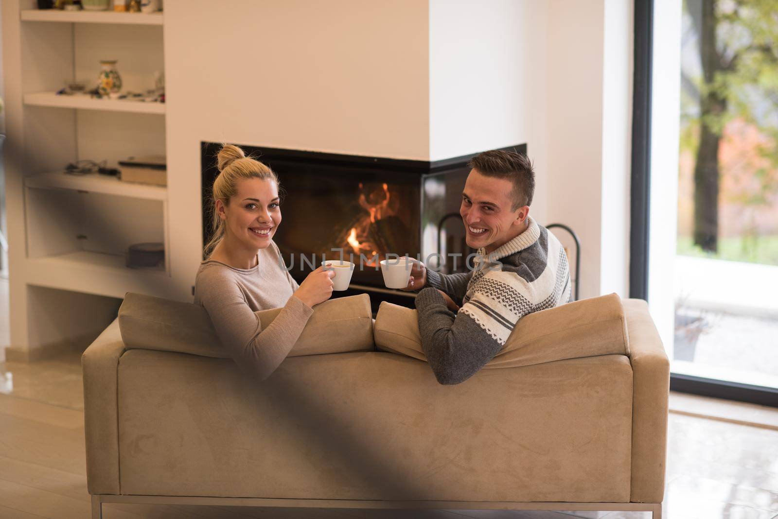 Young romantic couple sitting on sofa in front of fireplace at home, looking at each other, talking and drinking coffee at autumn day