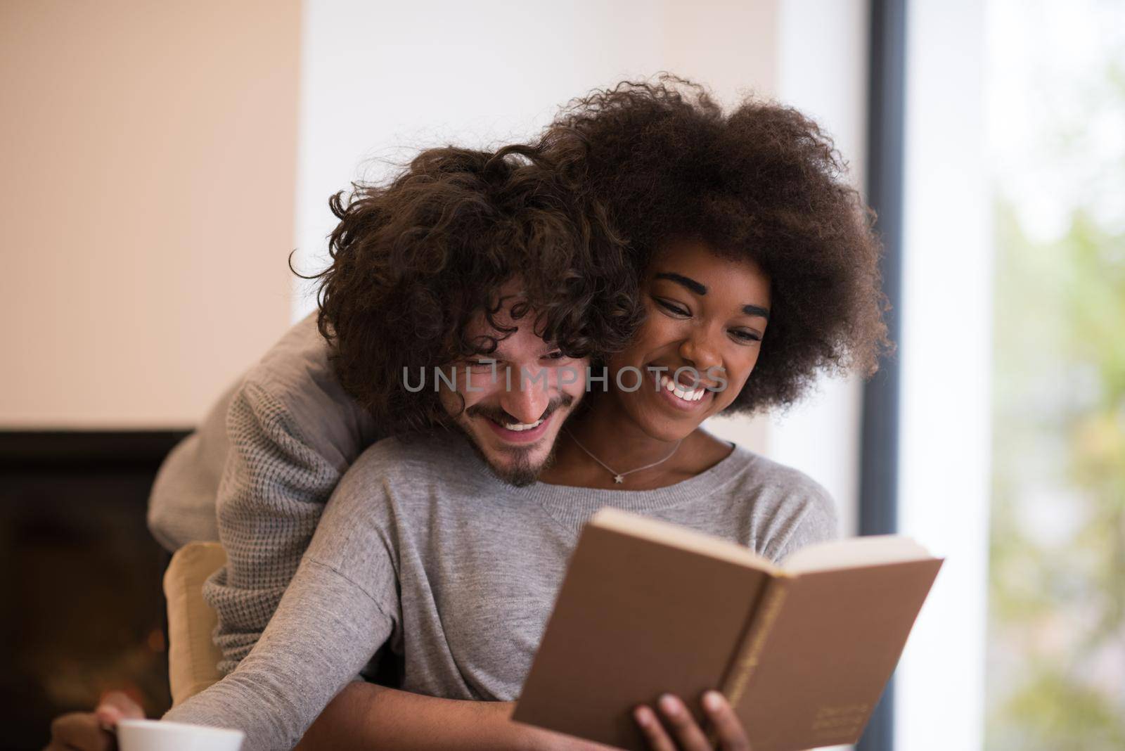 Young beautiful multiethnic couple hugging in front of fireplace at home when reading a book at autumn day