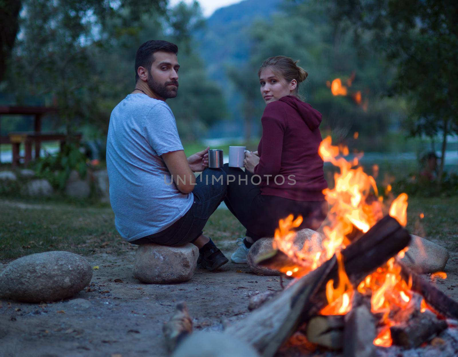 Young couple sitting around the campfire at evening holding a mugs of tea or coffee warming themselves