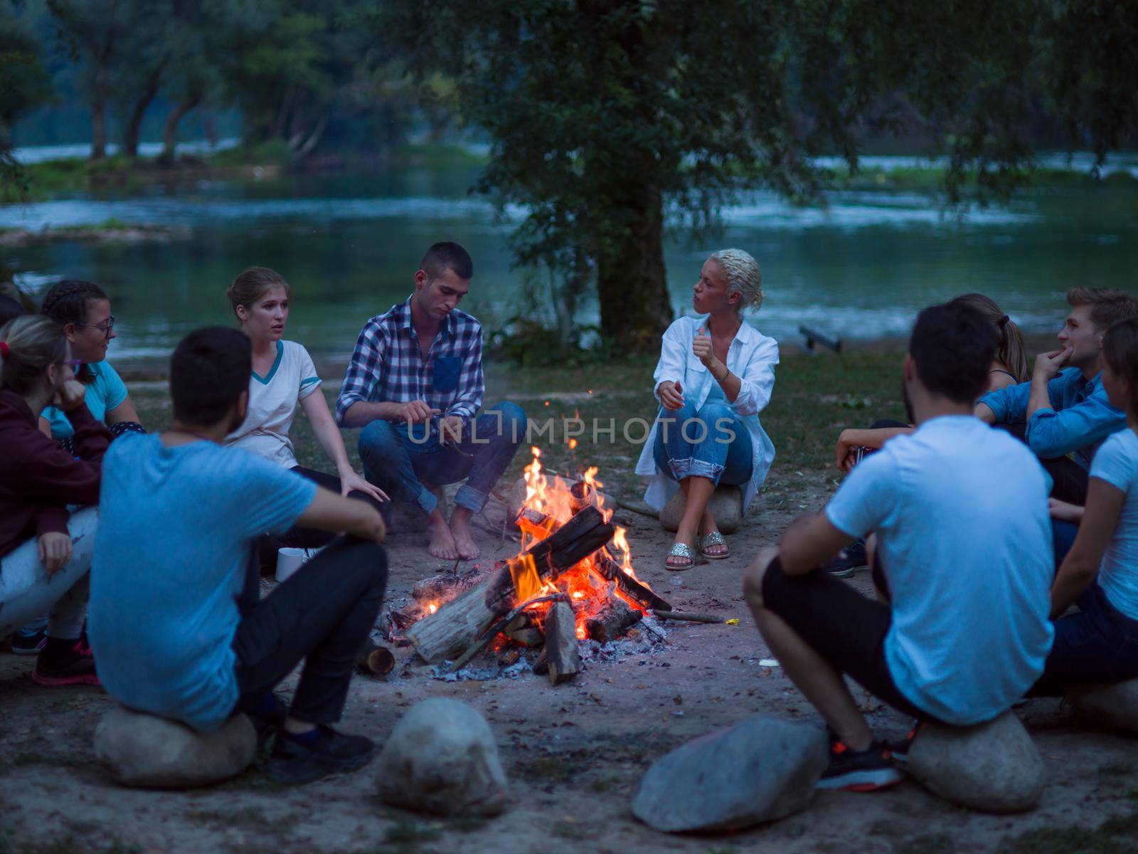 a group of happy young friends relaxing and enjoying  summer evening around campfire on the river bank