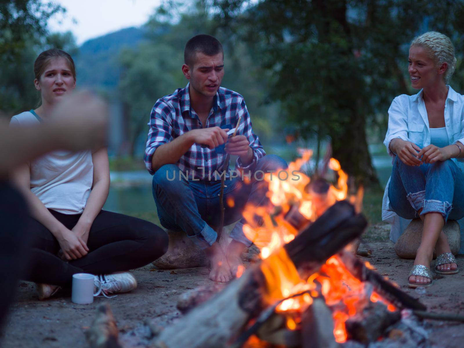 a group of happy young friends relaxing and enjoying  summer evening around campfire on the river bank