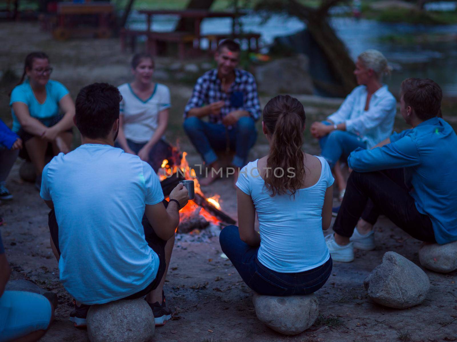 a group of happy young friends relaxing and enjoying  summer evening around campfire on the river bank