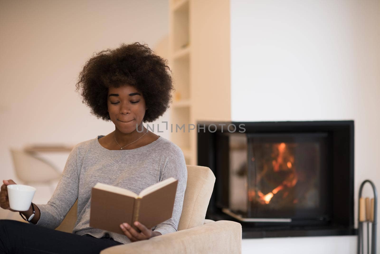 african american woman drinking cup of coffee reading book at fireplace. Young black girl with hot beverage relaxing heating warming up. autumn at home.