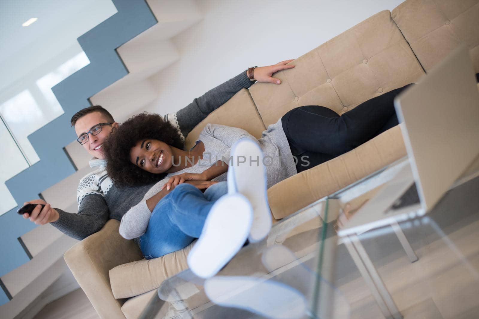 Young multiethnic couple relaxing on the sofa in the luxury living room, using a laptop and remote control