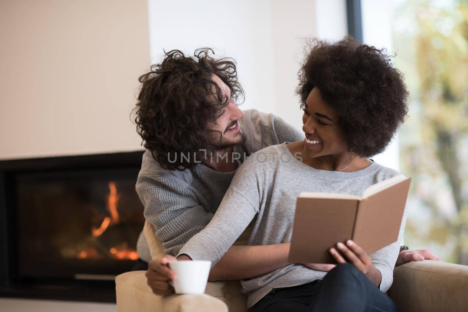 Young beautiful multiethnic couple hugging in front of fireplace at home when reading a book at autumn day