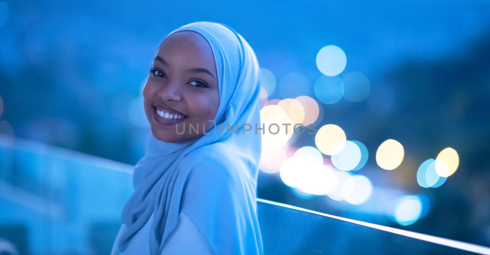 African  Muslim woman in night  at balcony  smiling at camera with city bokeh lights in background