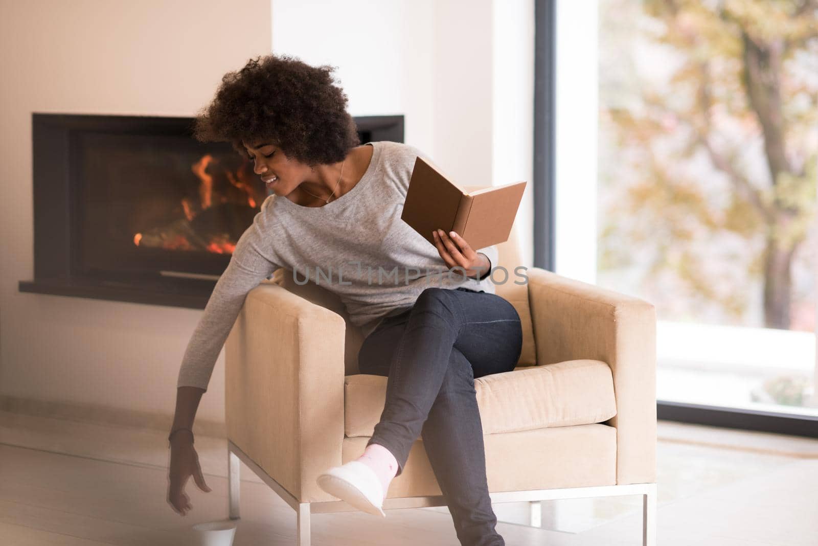 african american woman drinking cup of coffee reading book at fireplace. Young black girl with hot beverage relaxing heating warming up. autumn at home.