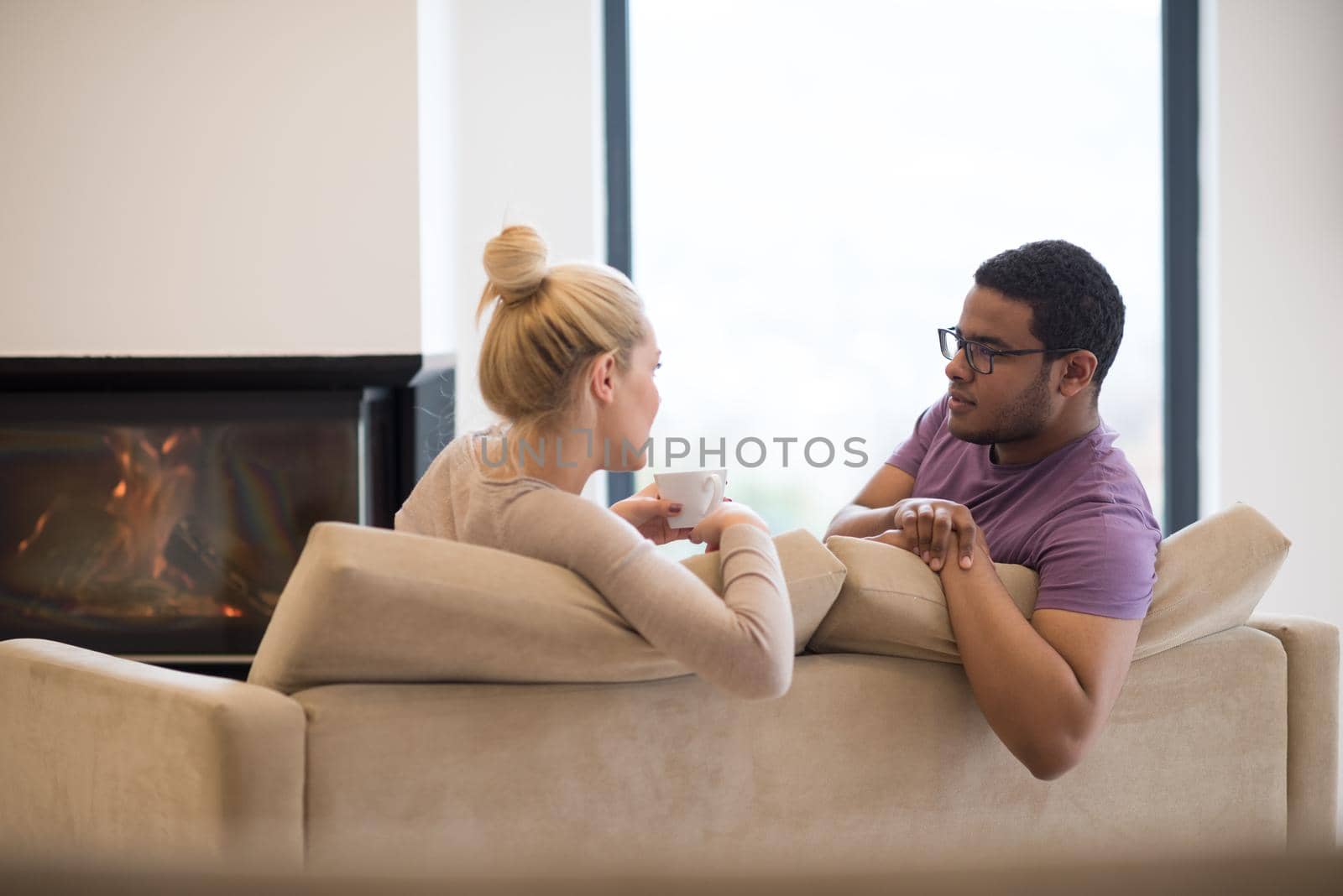 Young romantic multiethnic couple sitting on sofa in front of fireplace at home, looking at each other, talking and drinking coffee