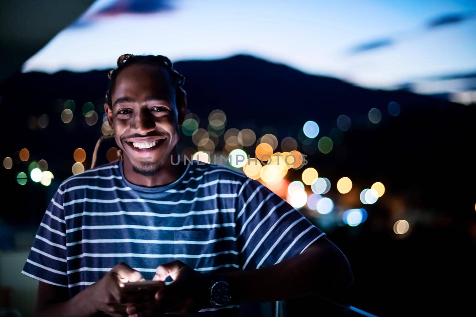 Young afro man on urban city street at night texting on smartphone with bokeh and neon city lights in background