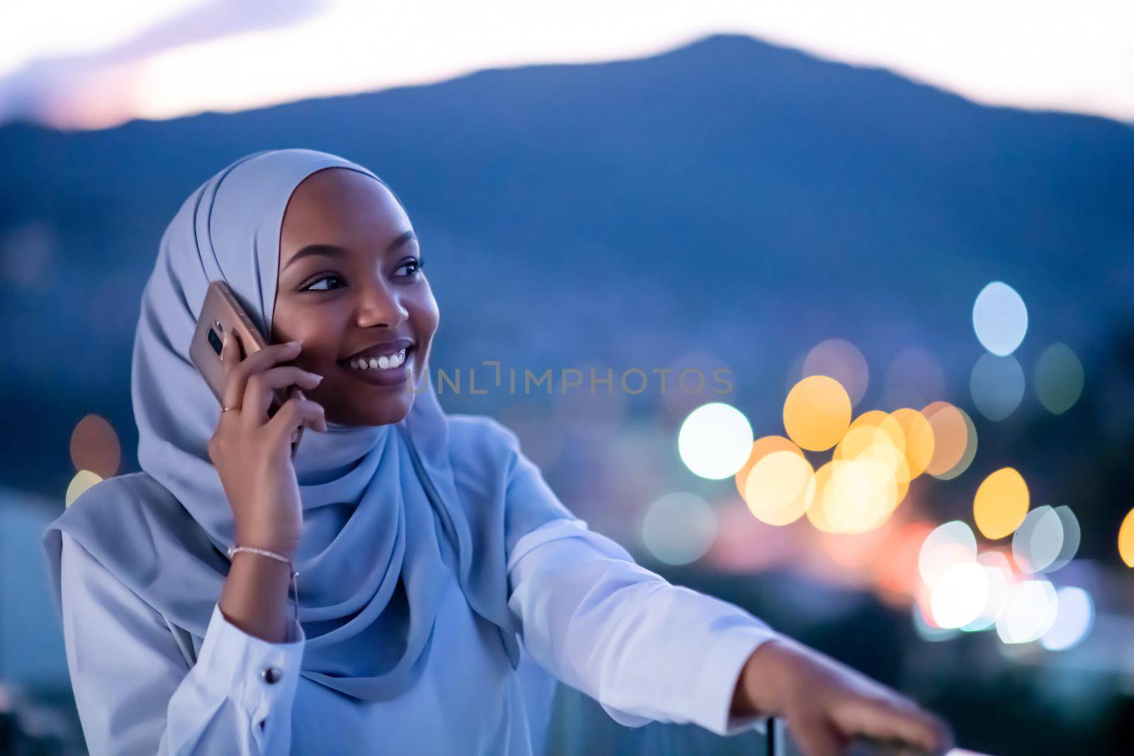 Young modern Muslim woman wearing scarf veil on urban city  street at night texting on smartphone with bokeh city light in background