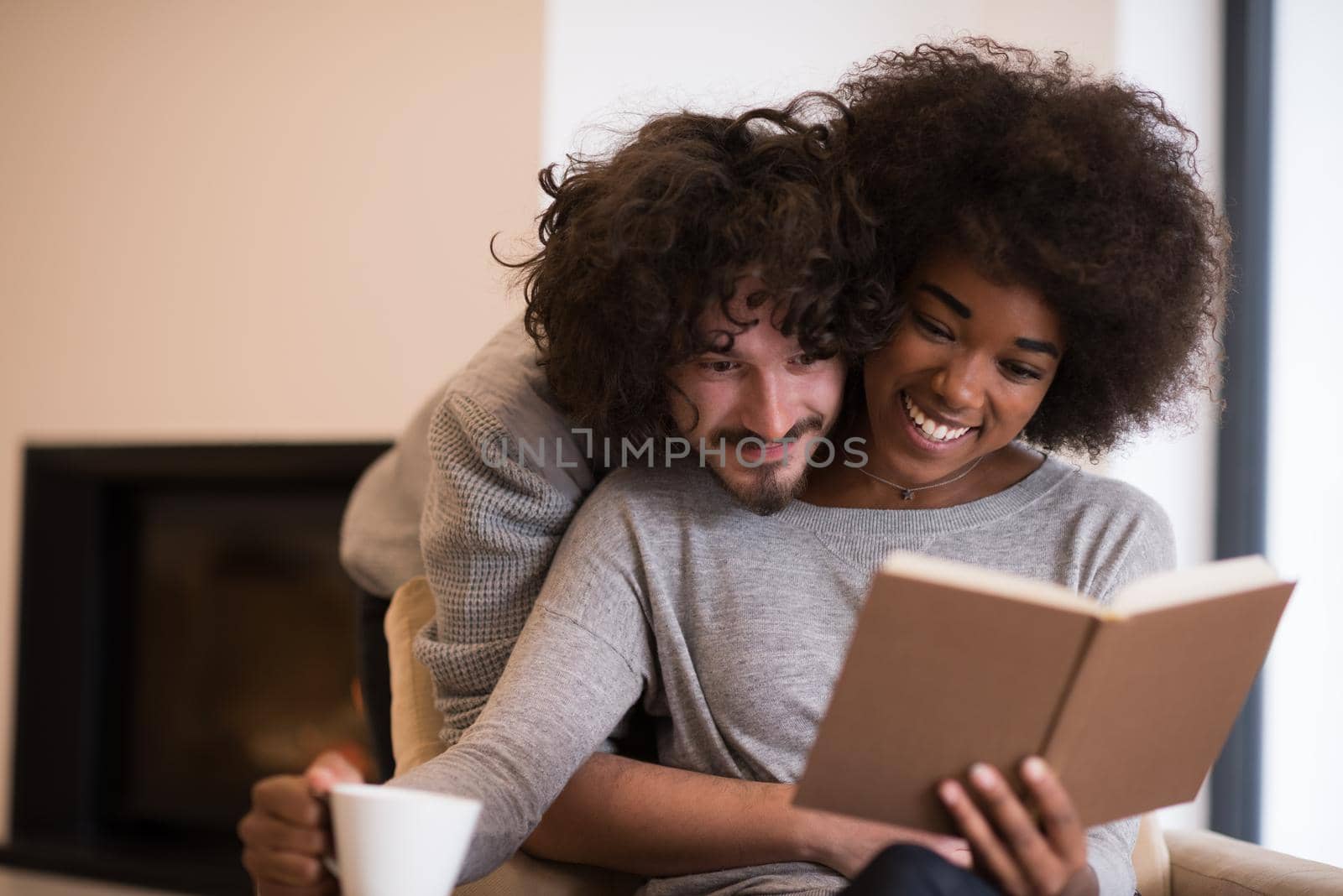 Young beautiful multiethnic couple hugging in front of fireplace at home when reading a book at autumn day