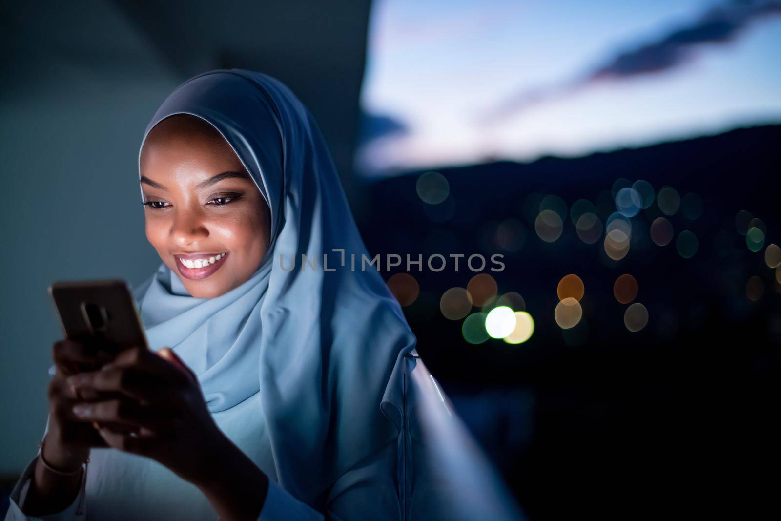 Young modern Muslim woman wearing scarf veil on urban city  street at night texting on smartphone with bokeh city light in background