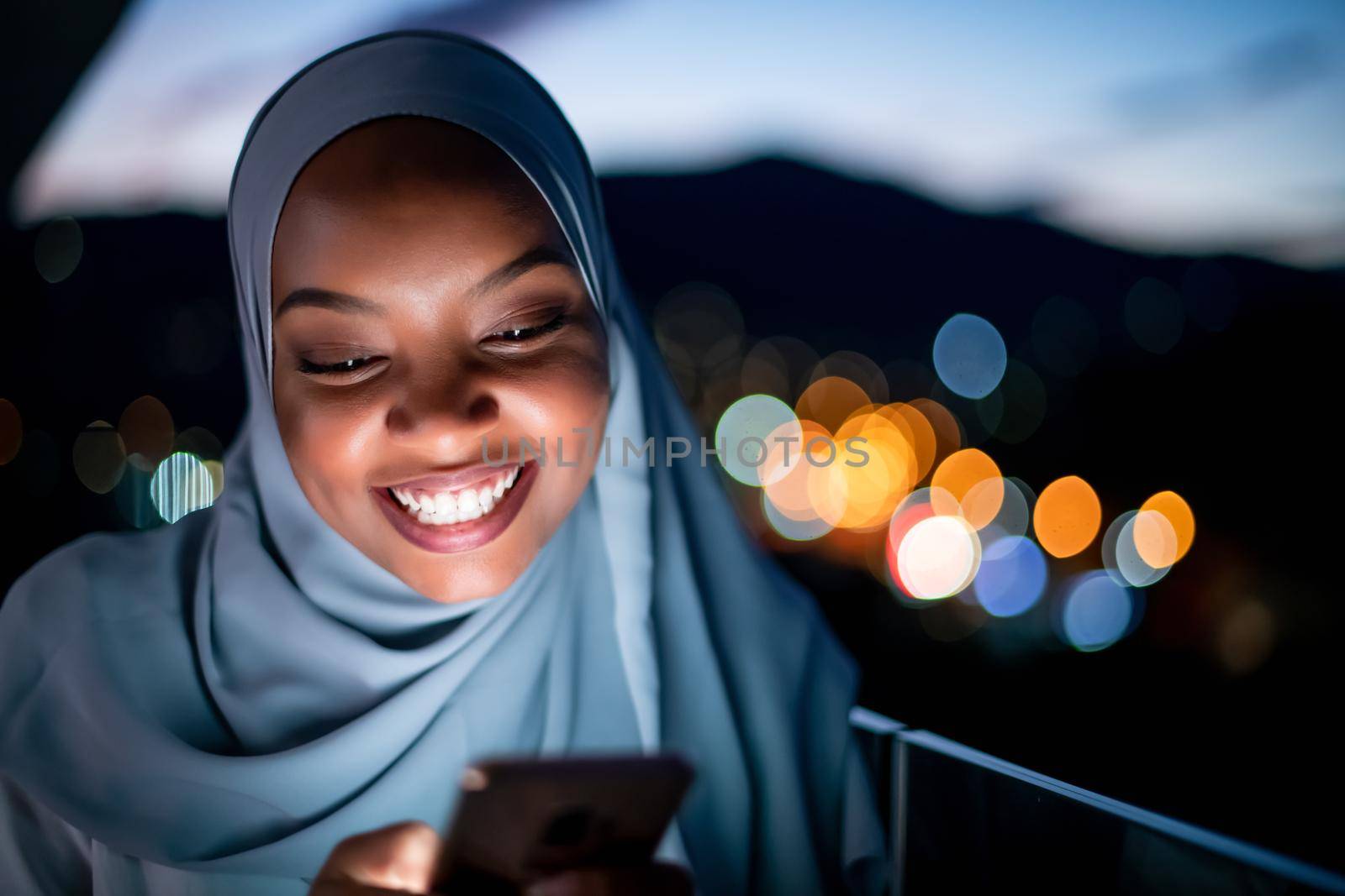 Young modern Muslim woman wearing scarf veil on urban city  street at night texting on smartphone with bokeh city light in background