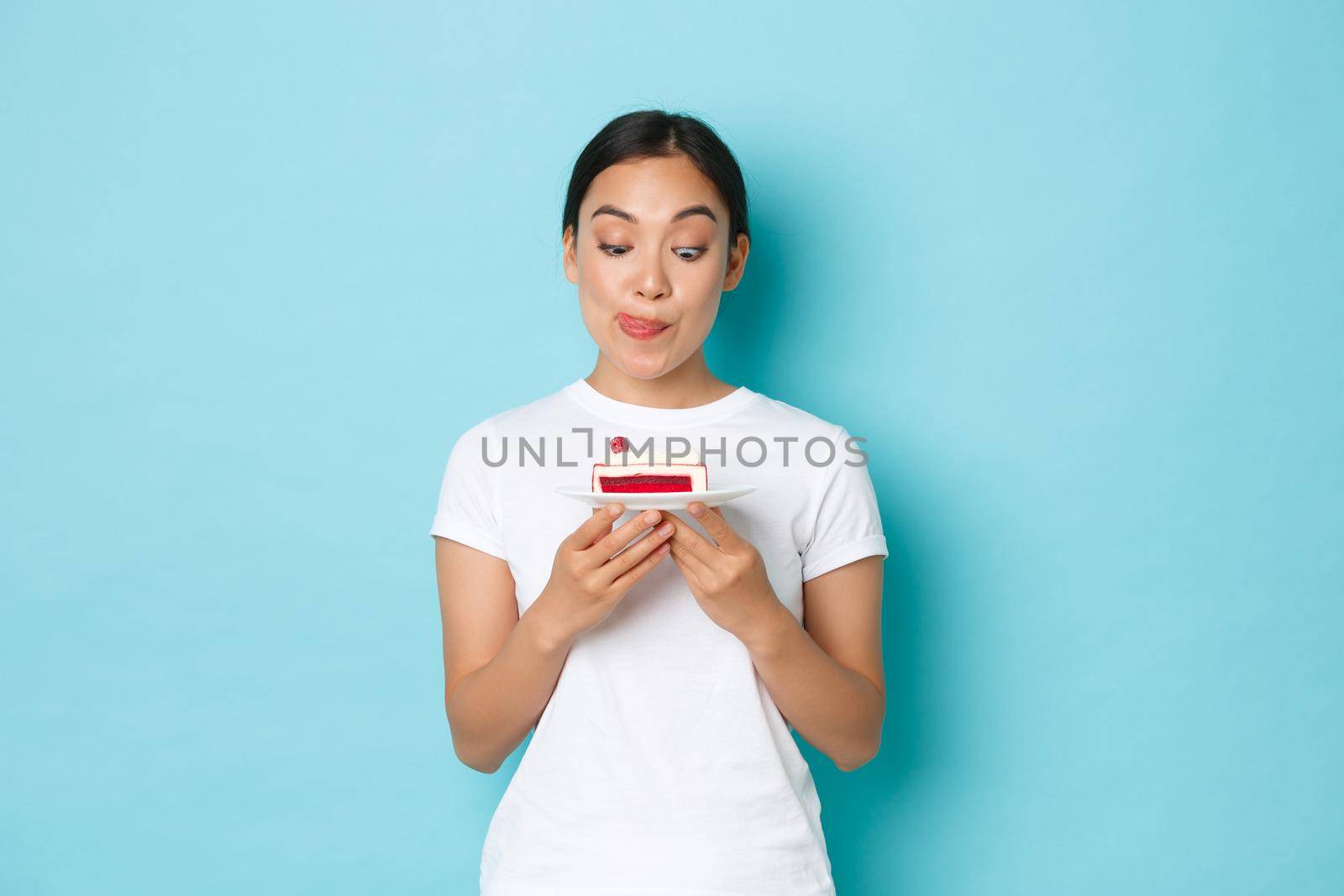 Holidays, lifestyle and celebration concept. Pretty and funny asian girl licking lips cant resist temptation while holding delicious piece of cake, want to eat dessert, standing light blue background.