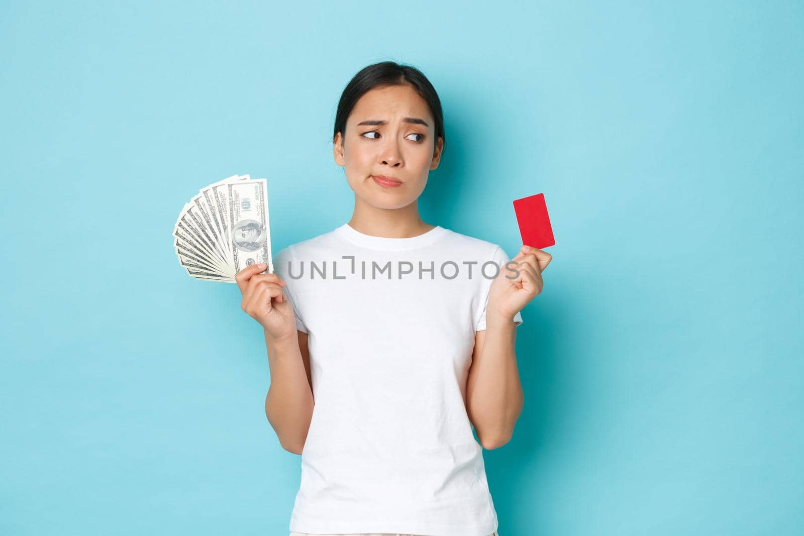 Shopping, money and finance concept. Indecisive and confused attractive asian girl cant decide whats better, credit card or cash, looking perplexed, standing thoughtful over blue background.