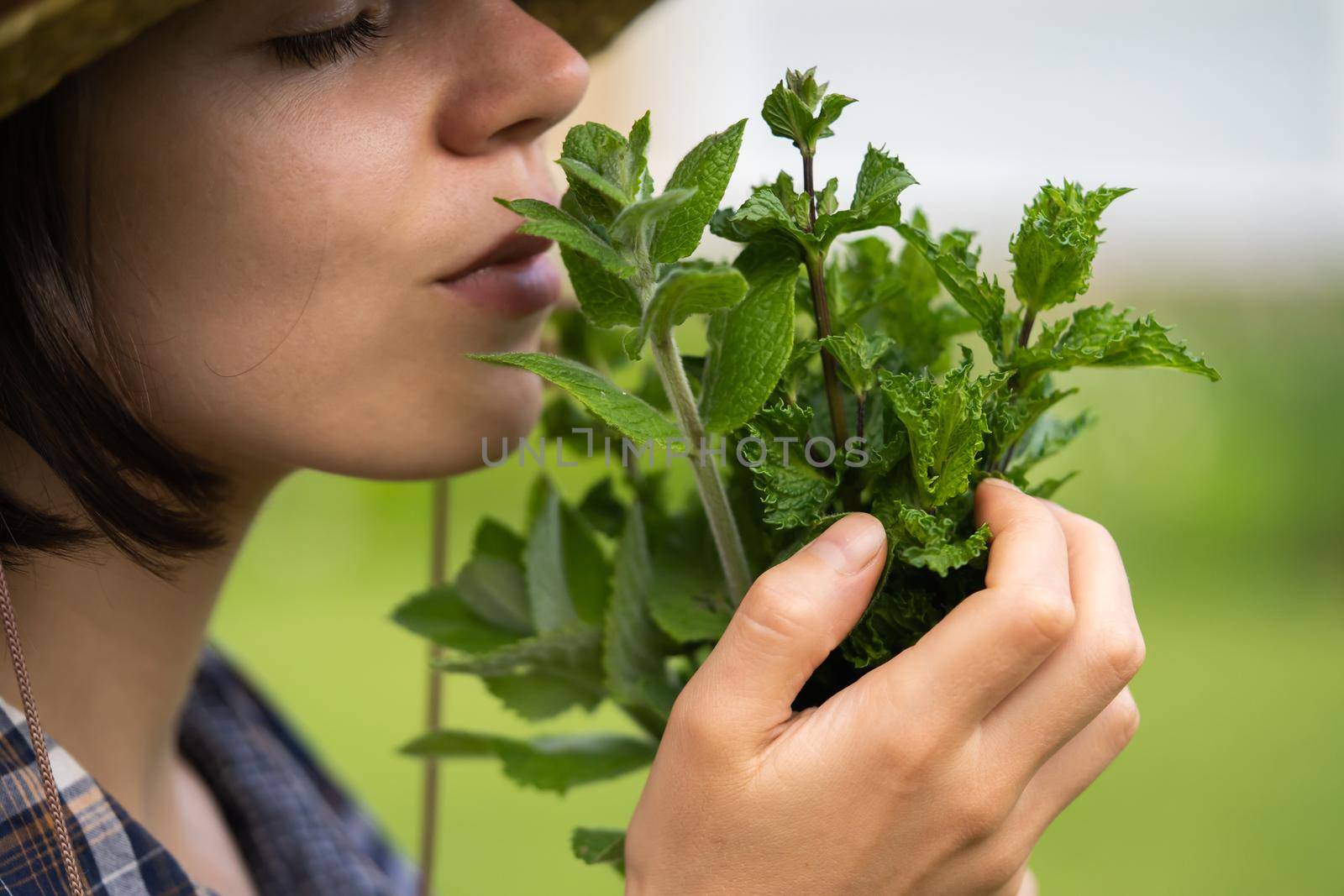 A young girl gardener in a straw hat holds a bouquet of harvested fresh mint and inhales its wonderful menthol scent, a woman is harvesting in the garden.