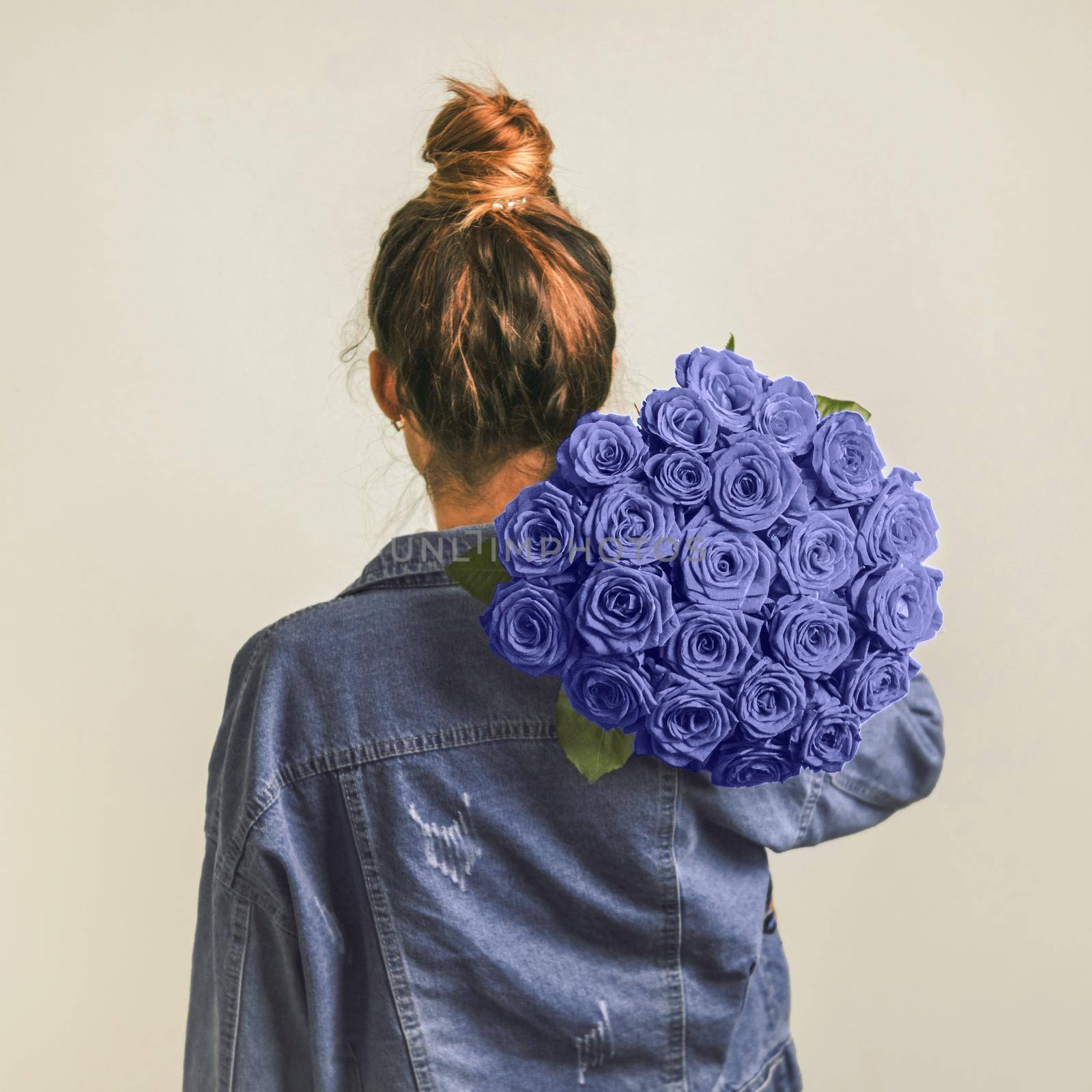 Back view of young woman in denim jacket holding bunch of violet roses on shoulder. Girl with bun updo in jeans holding flowers in very peri color 2022. Copy space. Demonstrating color of 2022 year