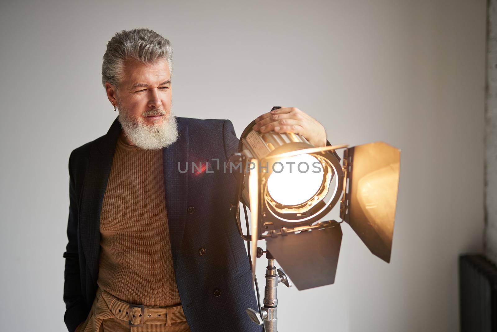 Portrait of handsome bearded middle aged man dressed elegantly standing next to studio spotlight while posing for camera over white background by friendsstock