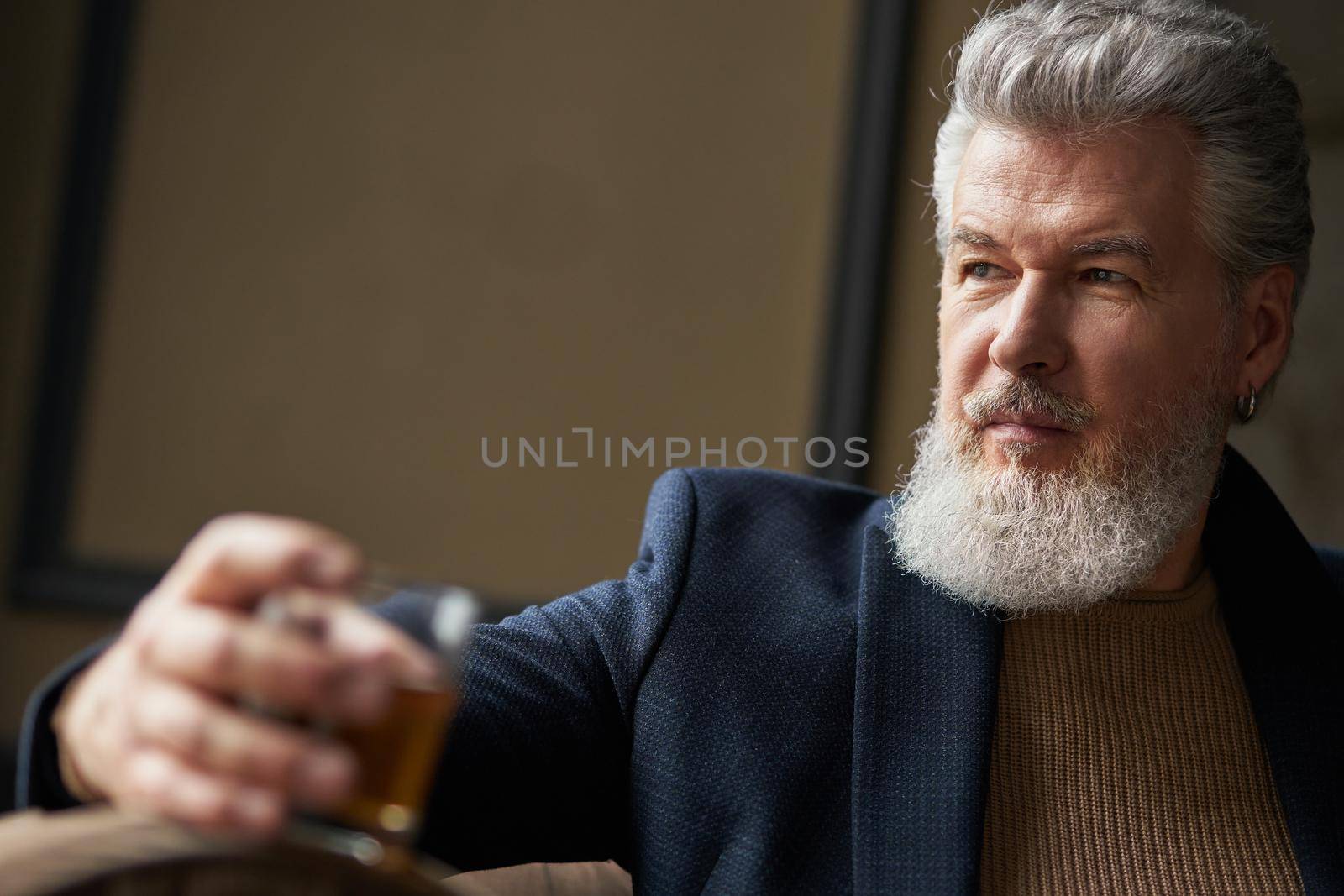 Confident middle aged business man holding glass of alcohol scotch whiskey in his hand while relaxing indoors. Focus on person. Lifestyle, success, people concept