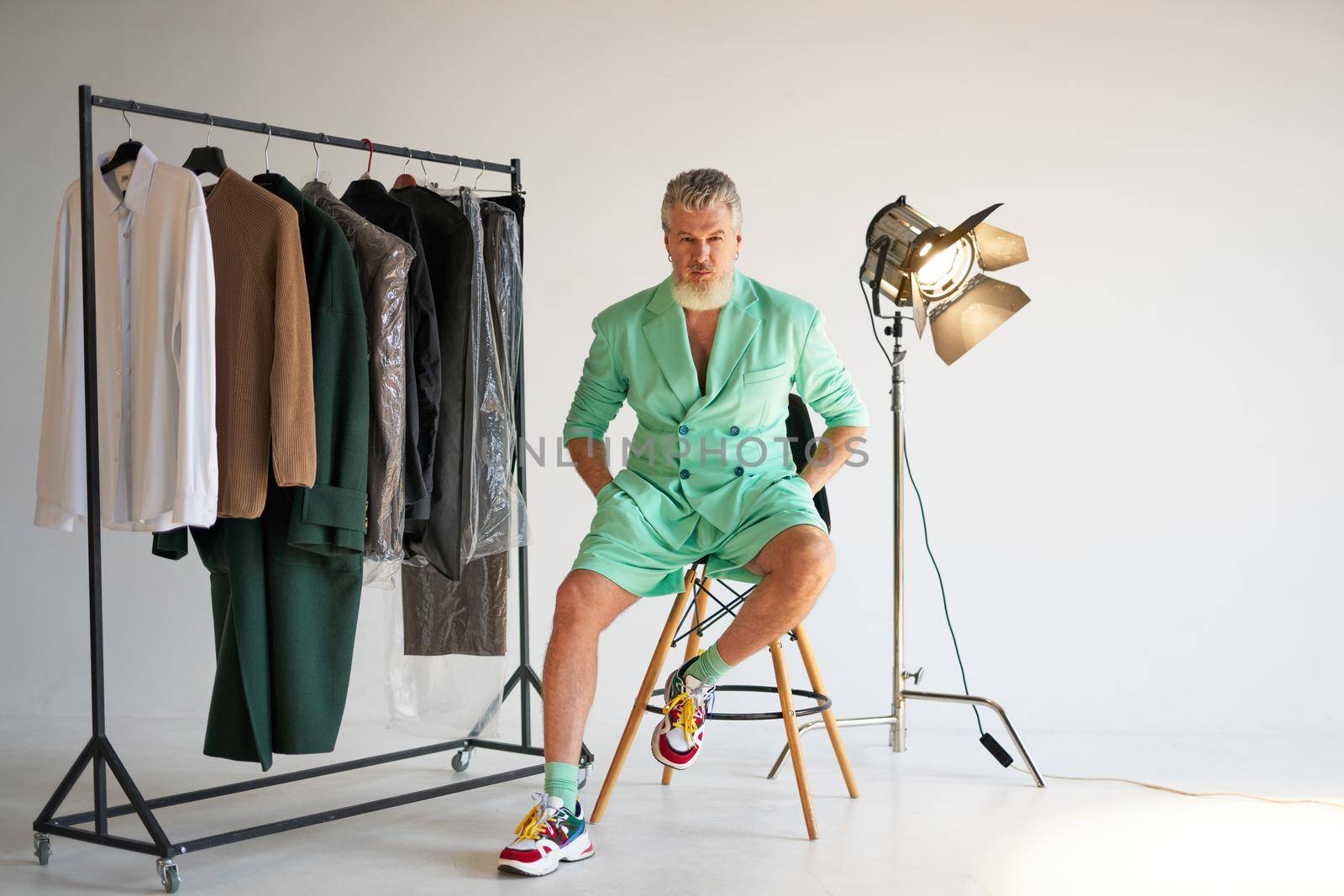 Studio shot of cool middle aged man with beard wearing stylish colorful outfit looking at camera while sitting next to clothes rail and studio spotlight over white background. Fashion photoshoot