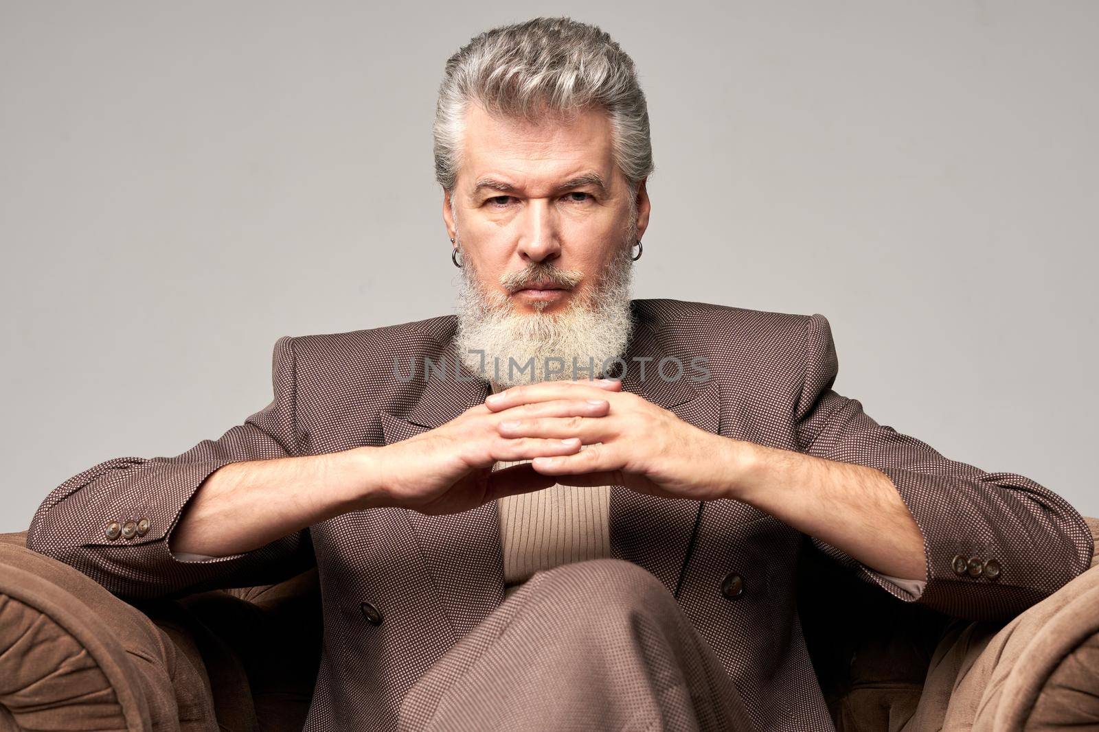 Portrait of brutal mature businessman with beard wearing elegant suit looking at camera, sitting with hands crossed in armchair while posing isolated over light background. Front view