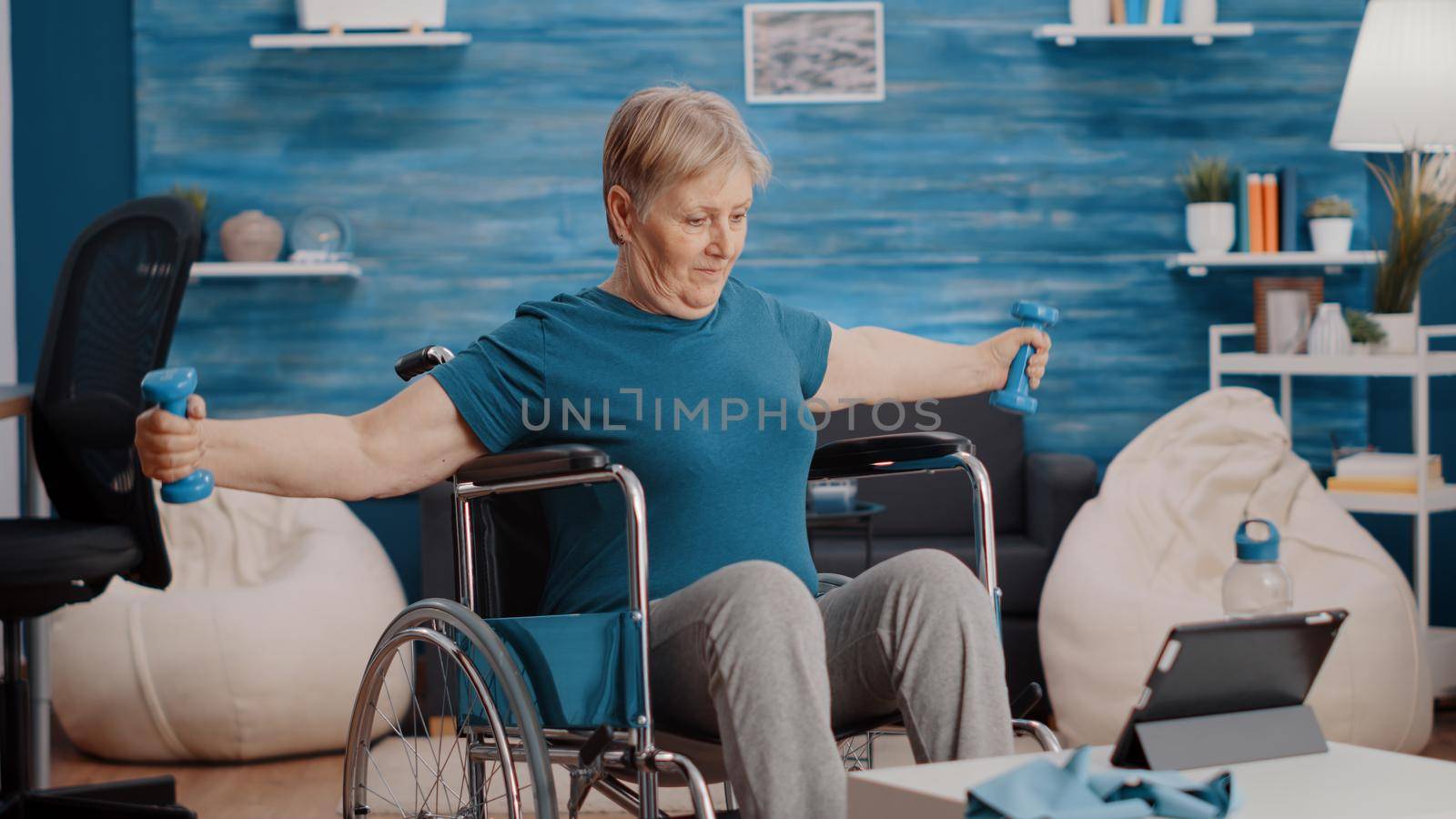 Senior adult sitting in wheelchair and doing exercise with weights while following training video on tablet. Elder woman with disability using dumbbells to train with online lesson.