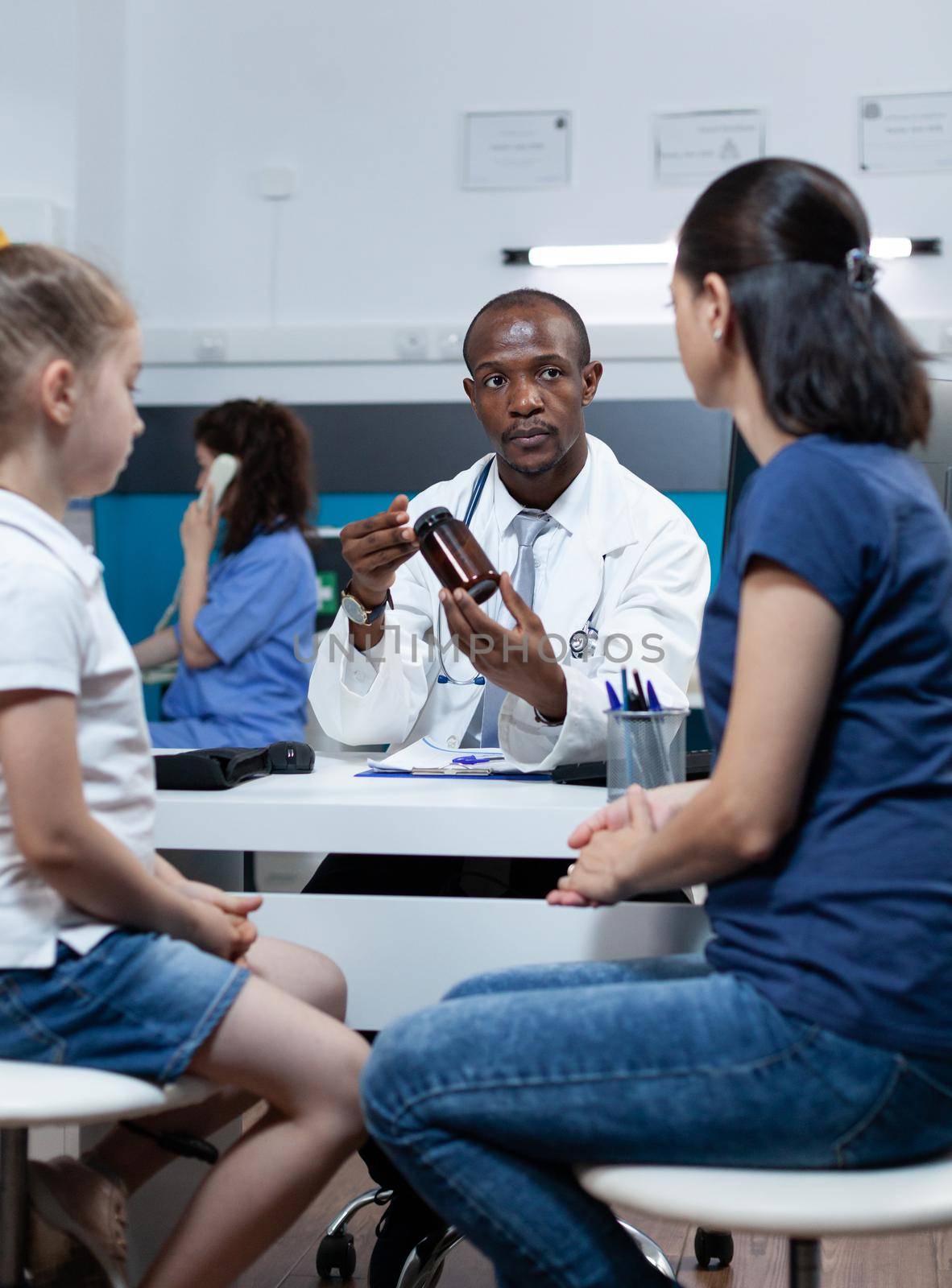 African american pediatrician doctor discussing sickness symptoms explaining medication treatment holding pills bottle during clinical examination in hospital office. Health care service