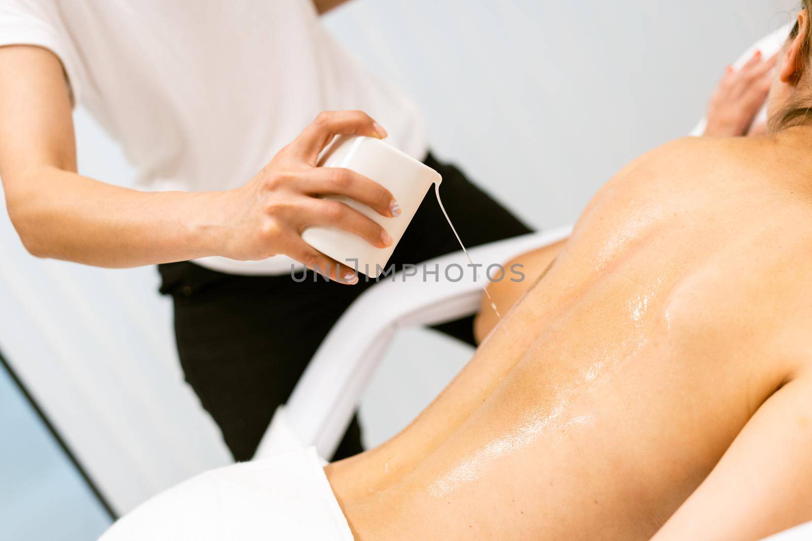 Beauty salon professional pouring oil from a massage candle on the back of his female patient.