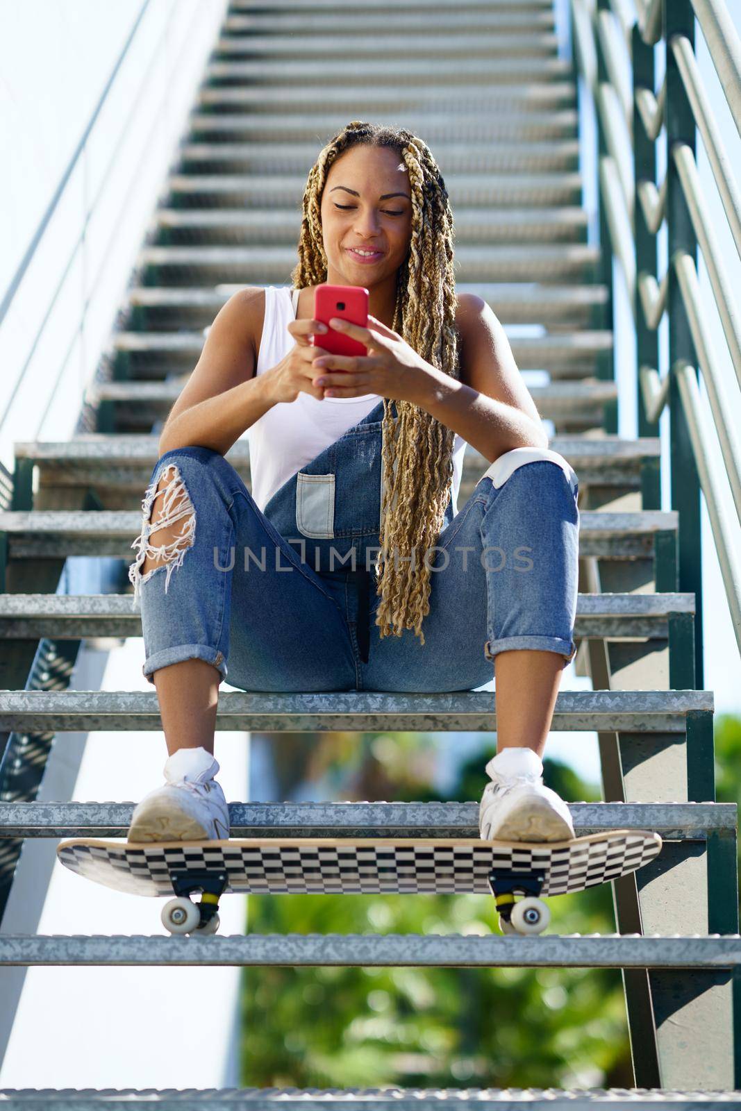 Young black woman with coloured braids, sitting on some steps while consulting her smartphone with her feet resting on a skateboard.