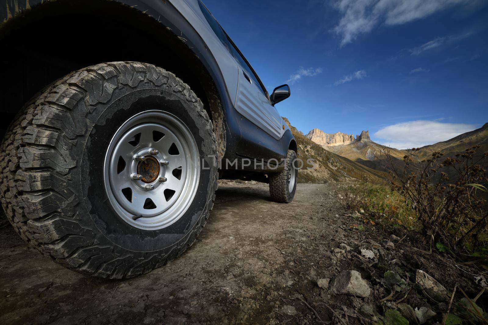 A large SUV car wheel with a large mud tread stands on a rocky road against the backdrop of mountains. Travel by off-road vehicles. Self-guided tourism by private car