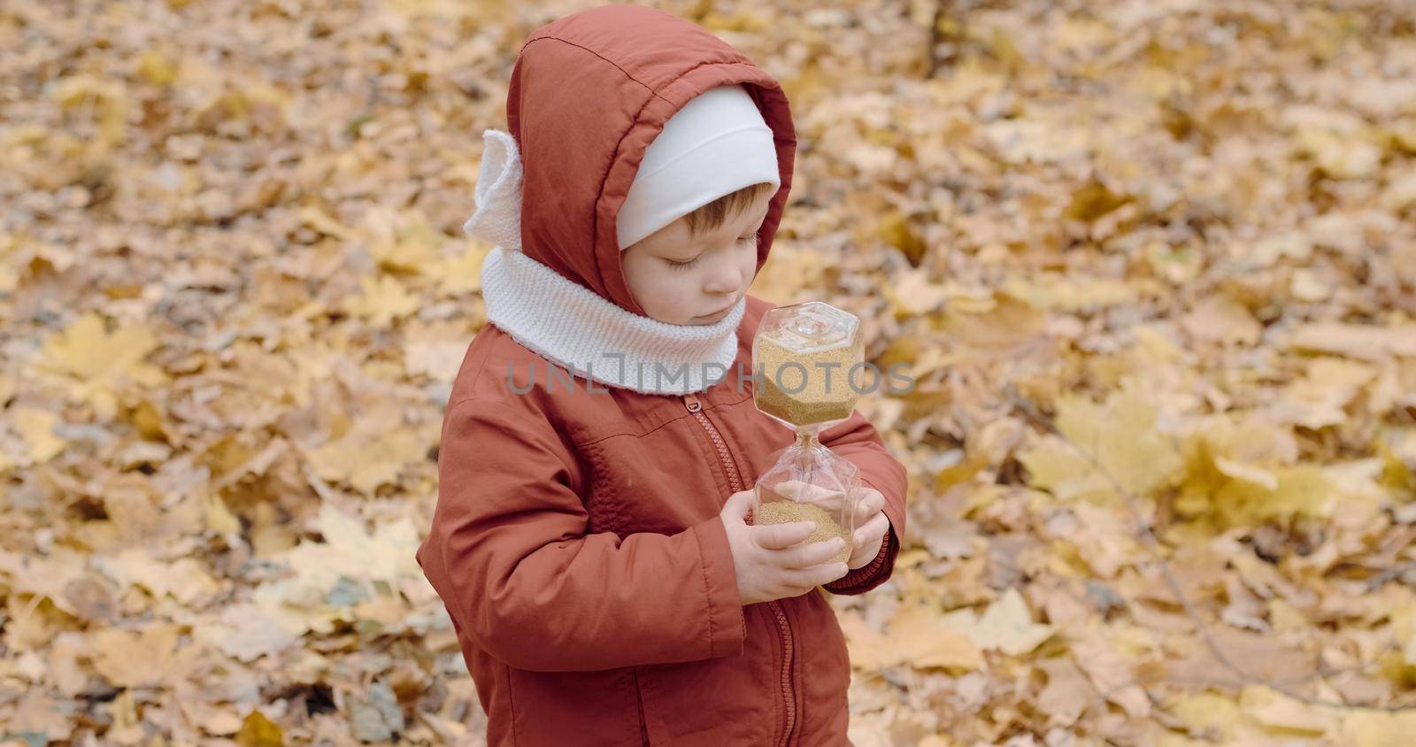 Little, a Caucasian girl is playing with a glass hourglass in the autumn forest. The concept of changing seasons, the transience of time and growing up