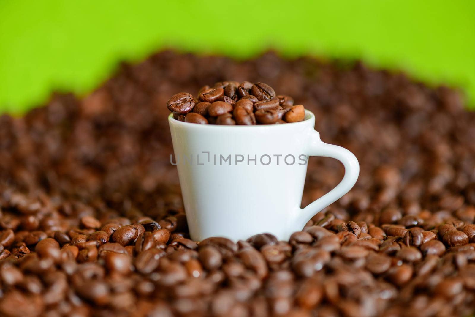 A white espresso cup filled with freshly roasted coffee beans on a table cluttered with coffee beans.