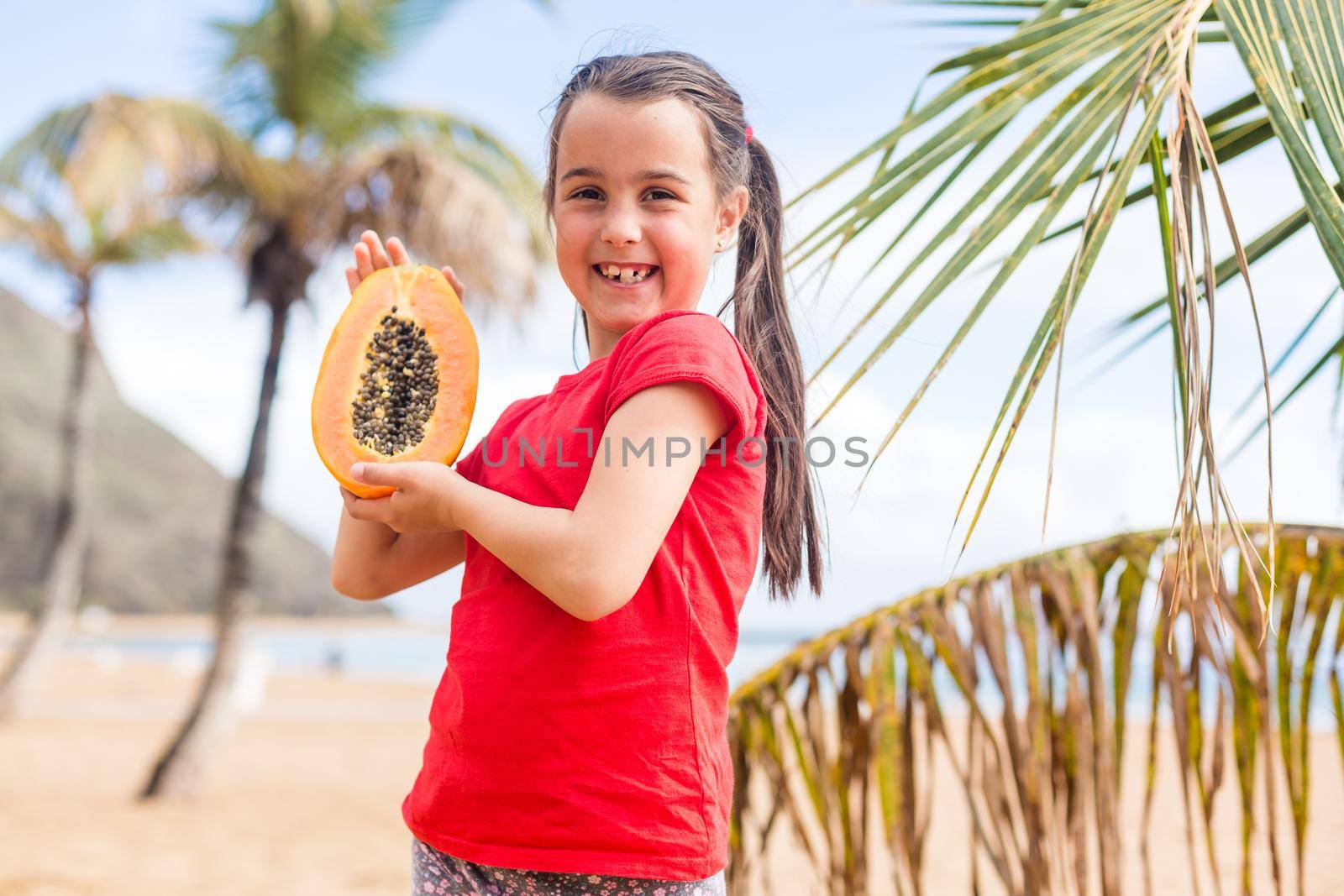 After playing on the sea black sand beach little cute girl picnics of natural vegetarian food - eating a orange slice of fresh ripe papaya, healthy children lifestyle in vacation on tropical island.