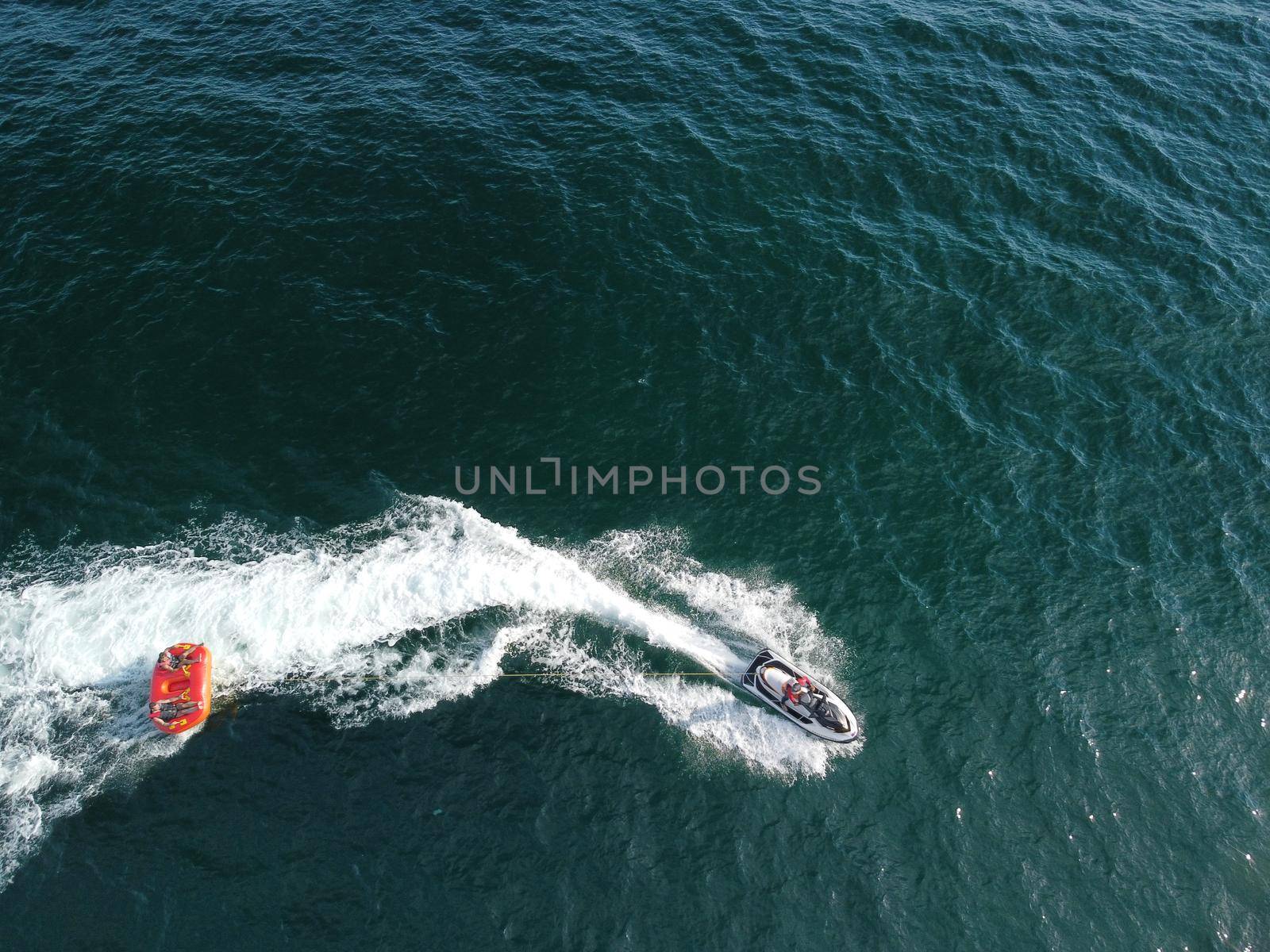 Happy people swim on air mattress behind a high speed water bike. Tourists ride on inflatable watercraft boat. Sea attraction. Water boat skating people on large Inflatable raft. Aerial copter view