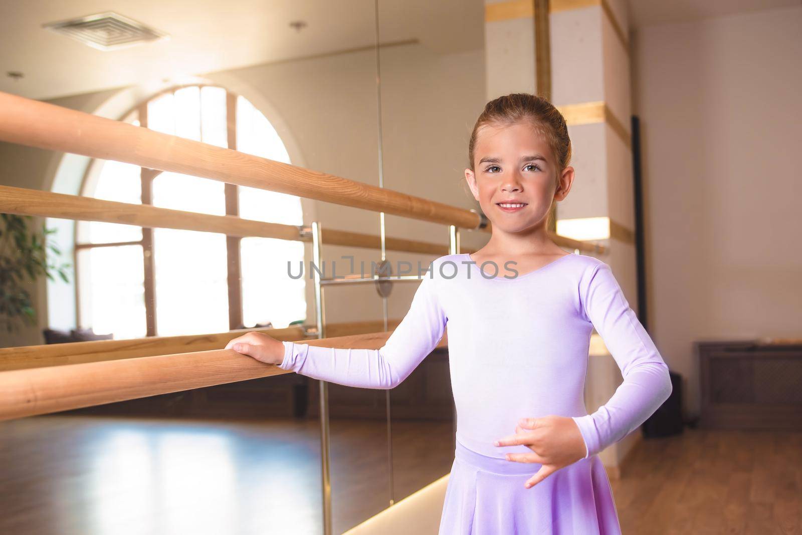 a little smiling girl in pointe shoes does stretching in ballet class near a frame and a big mirror by Nickstock