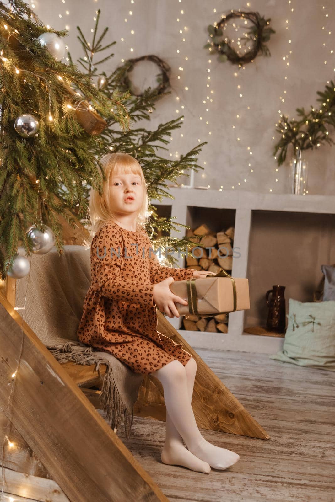 A little blonde girl is sitting on a wooden staircase in a Scandinavian interior decorated in a New Year's style. A child holds a Christmas gift in a craft package.