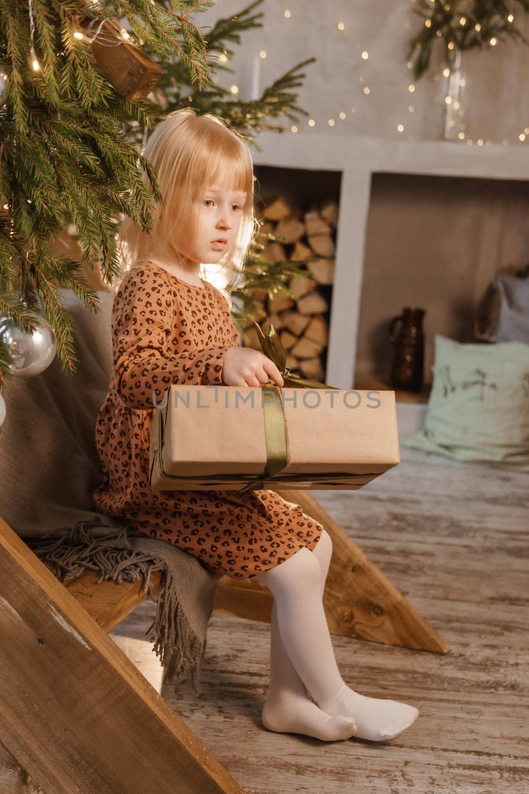 A little blonde girl is sitting on a wooden staircase in a Scandinavian interior decorated in a New Year's style. A child holds a Christmas gift in a craft package.