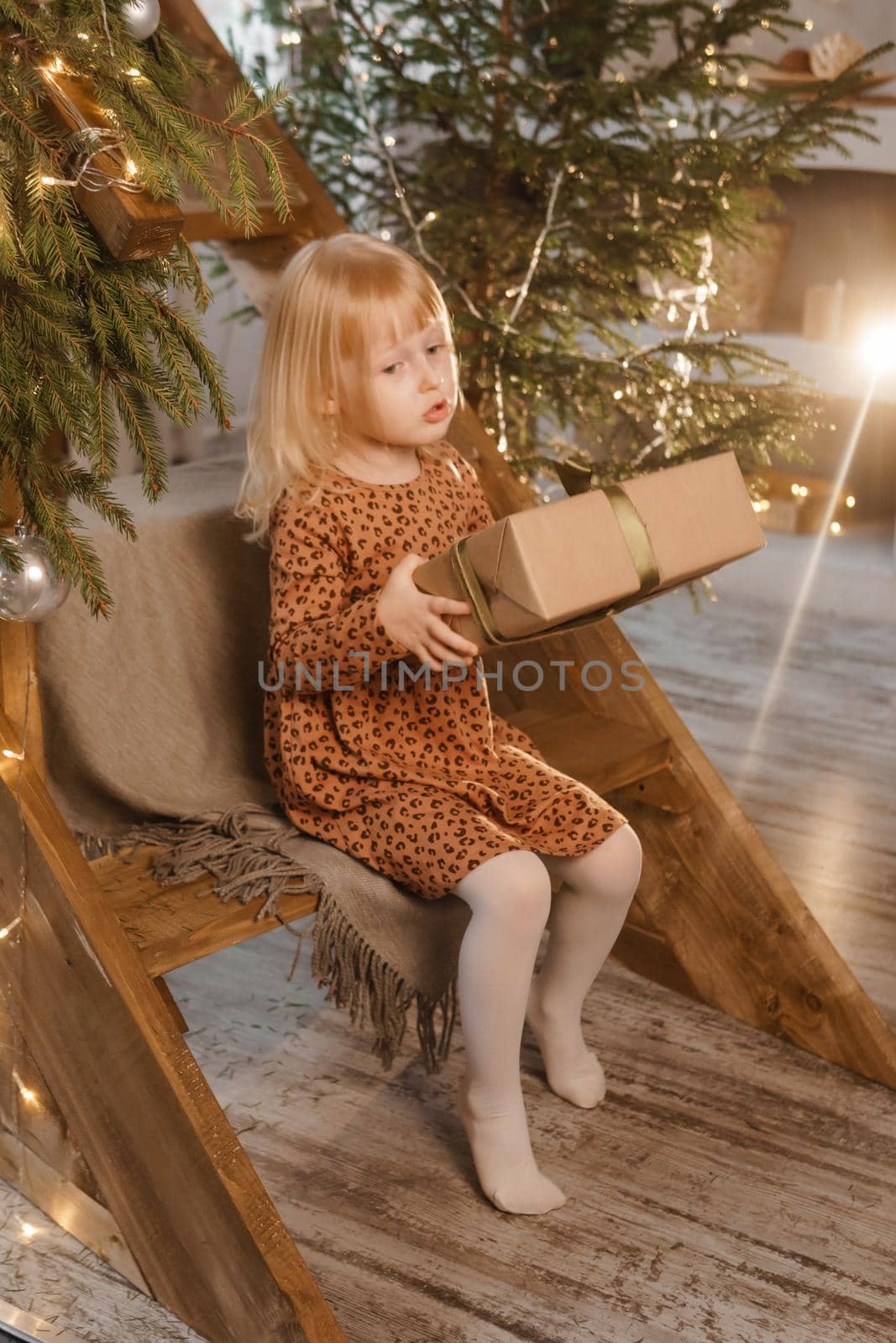 A little blonde girl is sitting on a wooden staircase in a Scandinavian interior decorated in a New Year's style. A child holds a Christmas gift in a craft package.