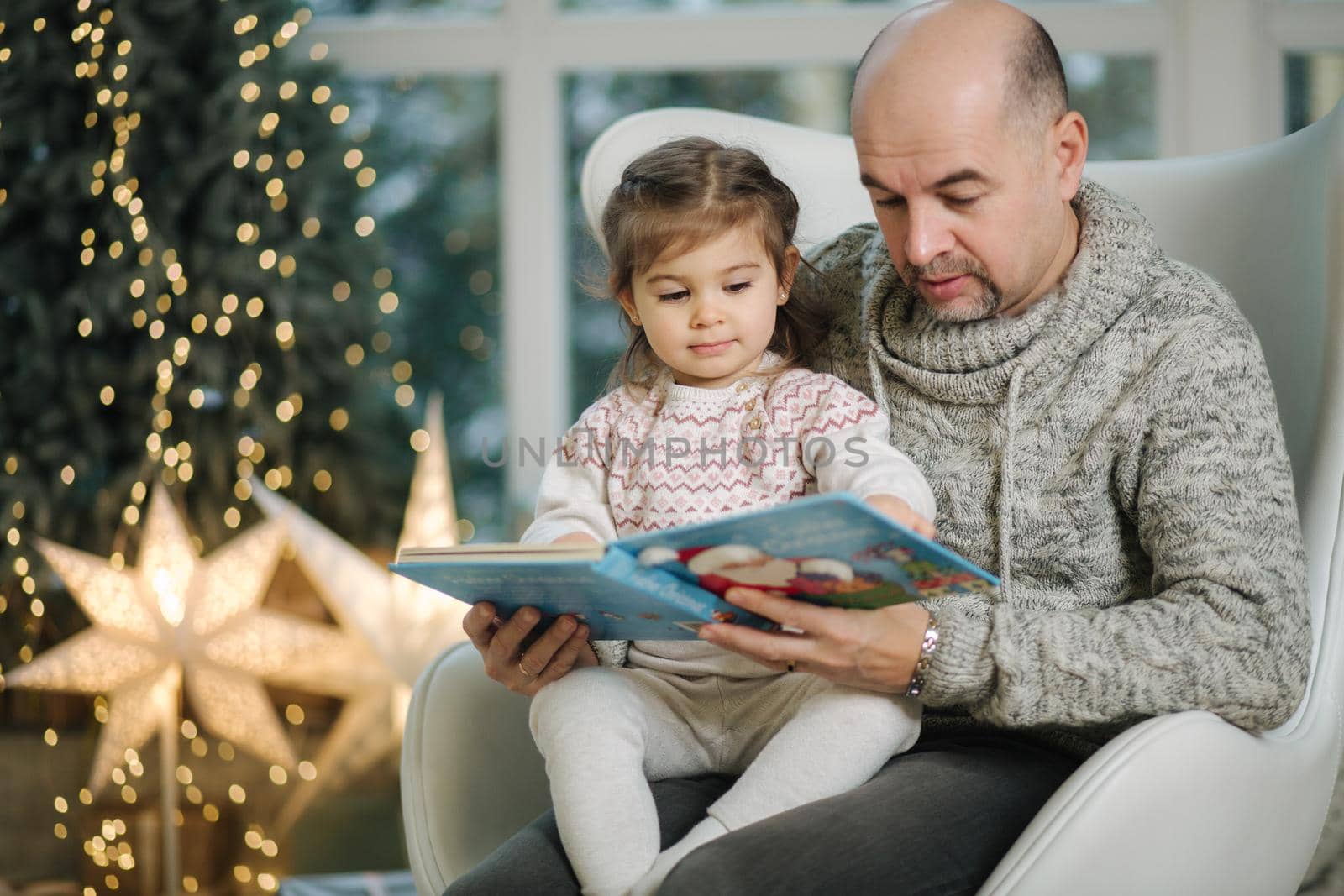 Young grandfather play and read book with her adorable grandaugher nea fir tree. Christmas mood.