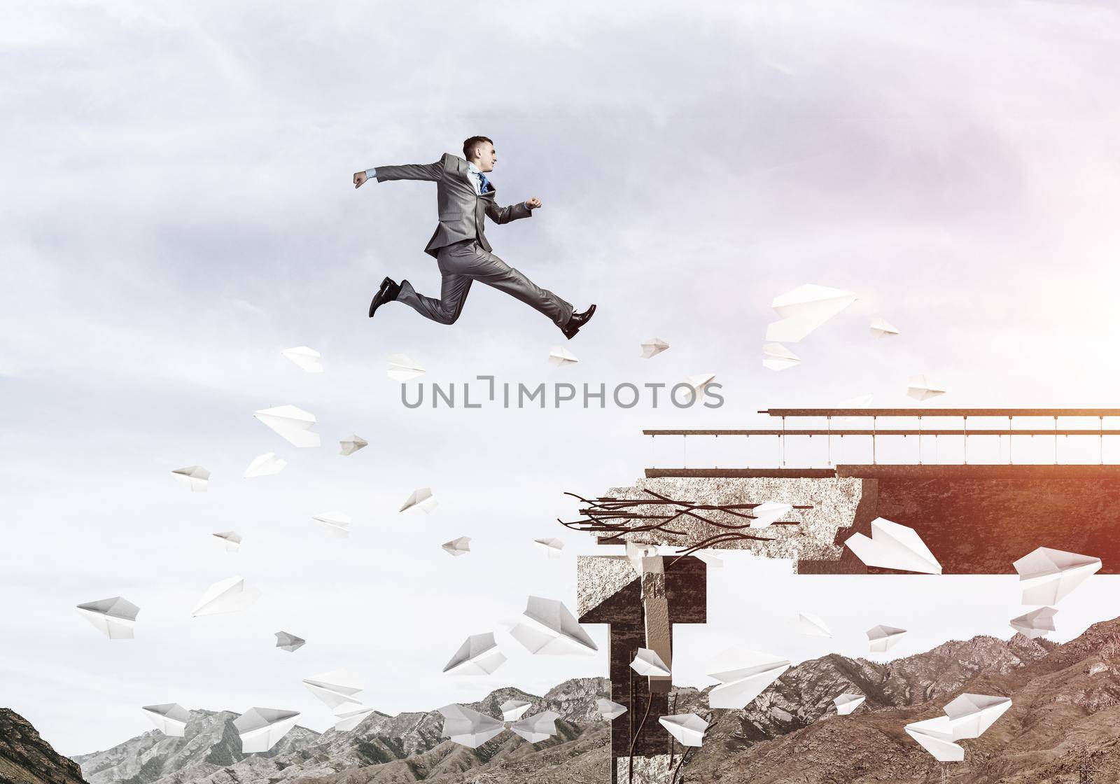 Businessman jumping over gap in bridge among flying paper planes as symbol of overcoming challenges. Skyscape with sunlight and nature view on background. 3D rendering.