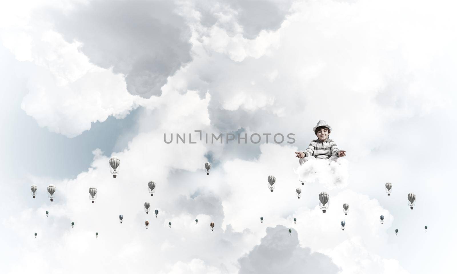 Young little boy keeping eyes closed and looking concentrated while meditating among flying balloons in the air with cloudy skyscape on background.